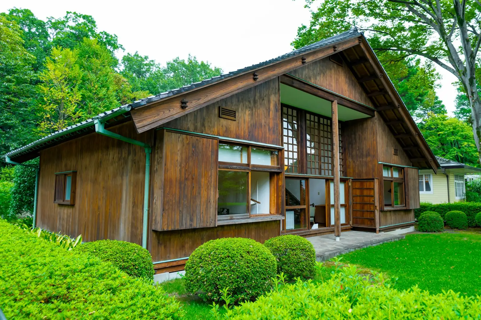 A small wooden house with a big window, set back on a green lawn with trimmed shrubs